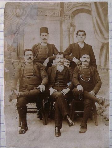 " Φωτογραφία με τους κατοίκους του χωριού. Στη φωτογραφία οι τρεις από αυτούς φοράνε τις παραδοσιακές φορεσιές του τόπου " Ομαδική φωτογραφία πέντε αντρών . Δυο αντρές στέκονται όρθιοι ακριβώς πίσω από τρεις καθιστούς άντρες. Οι δυο άντρες που βρίσκονται στην αριστερή πλευρά της φωτογραφίας φορούν πουτούρια ,πουκαμίσα ,αμπά και τσαρούχια. Στο κέντρο καθιστός είναι ένας άντρας με κουστούμι .Στην δεξιά πλευρά ο άντρας που είναι όρθιος φοραεί κουστούμι , ενω αυτος που καθεται φορά την παραδοσιακή ενδυμασία.