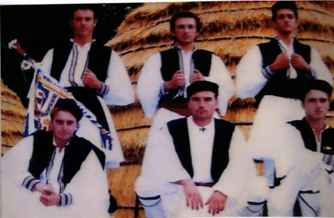 Φωτογραφία νεαρών αντρών με το παραδοσιακό σαρακατσάνικο ένδυμα. Το ένδυμα αποτελείται από το στραγγάνι(μαυρο στρογγυλό καπέλο),  το πουκάμισο, το τσιπκένι (γιλεκο), το ζωστάρι , την φουστανέλα και τις κάλτσες. Οι νεαροι φωτογραφίζονται έξω από μια καλύβα. Έγχρωμη φωτογραφία
